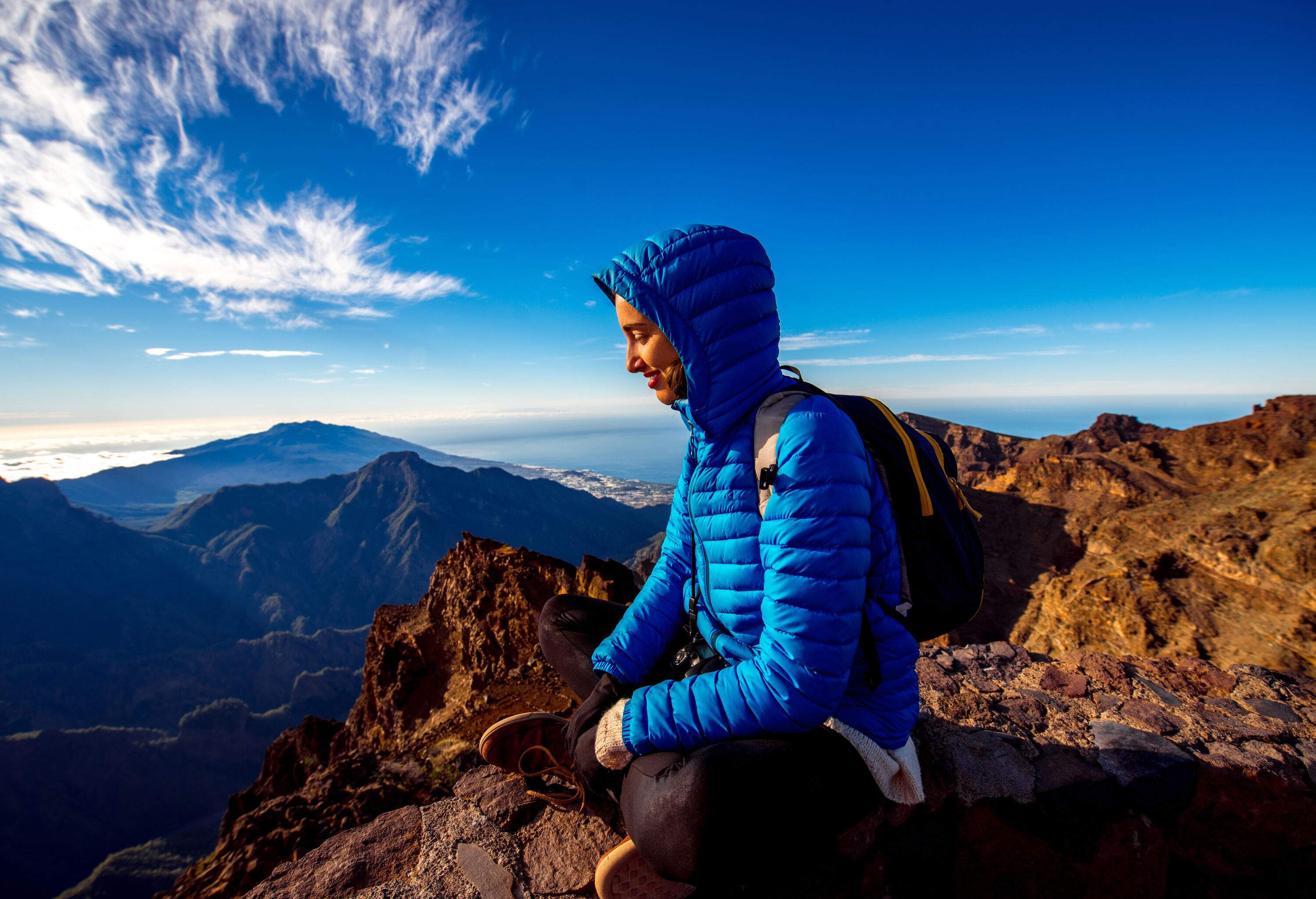 A smiling person in a blue jacket sits cross-legged on a cliff's edge and looks down at the scenery below.