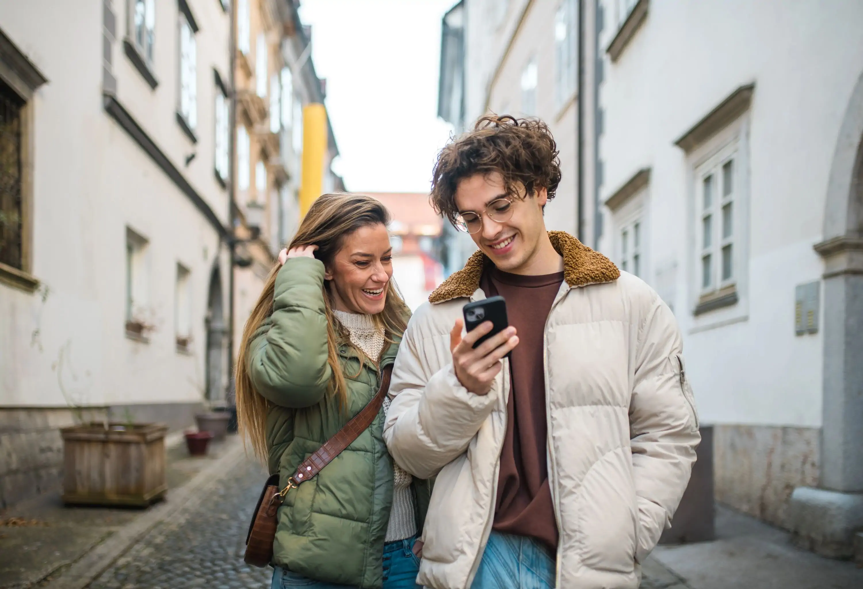 Happy Caucasian woman with her young adult son standing in an urban setting engaging in a video call. Both wear casual autumn jackets, sharing a joyful moment with their mobile device.