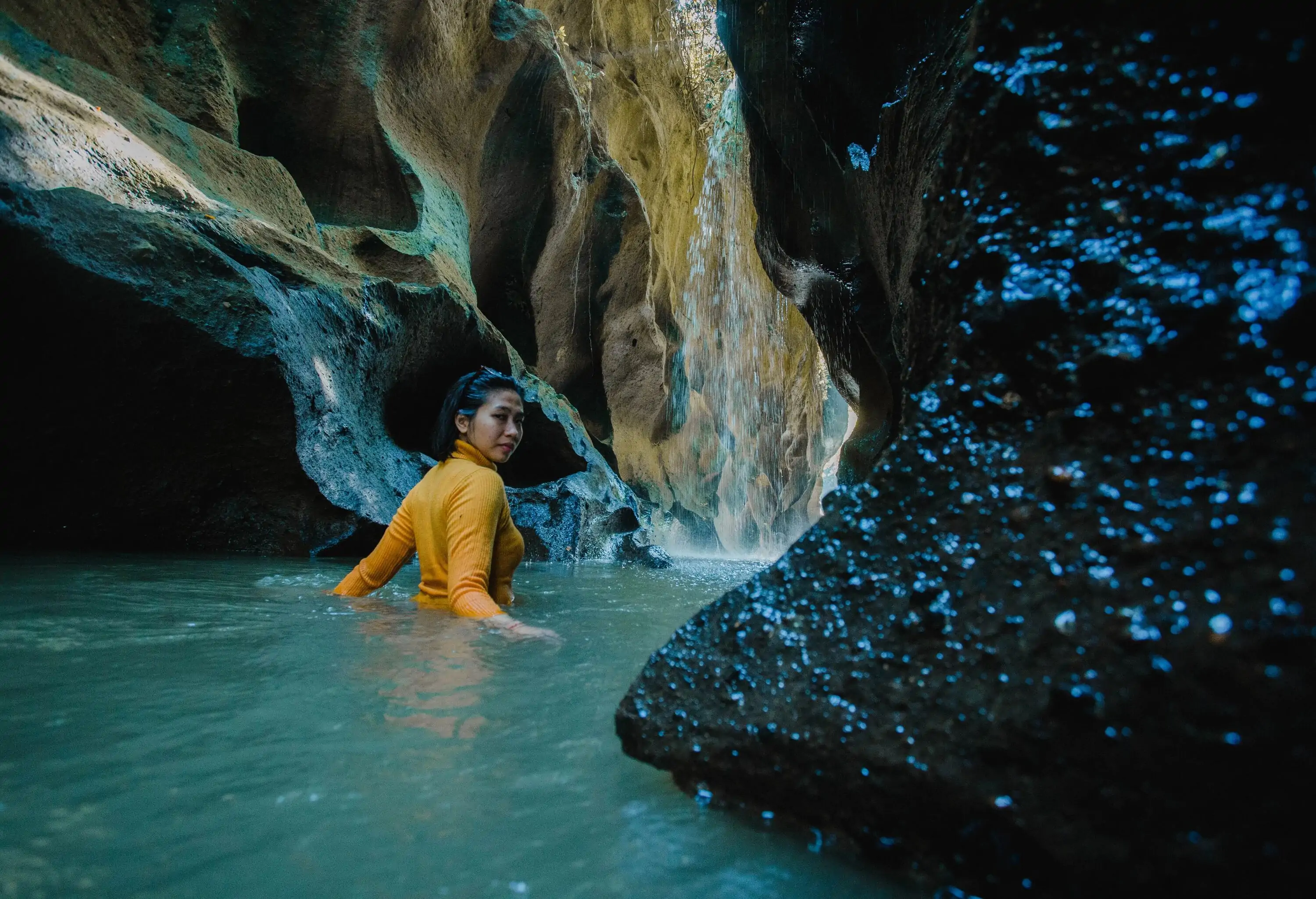 A person in a dark yellow sleeved turtleneck top relaxes in a natural pool buried between the gaps of towering steep rocks.