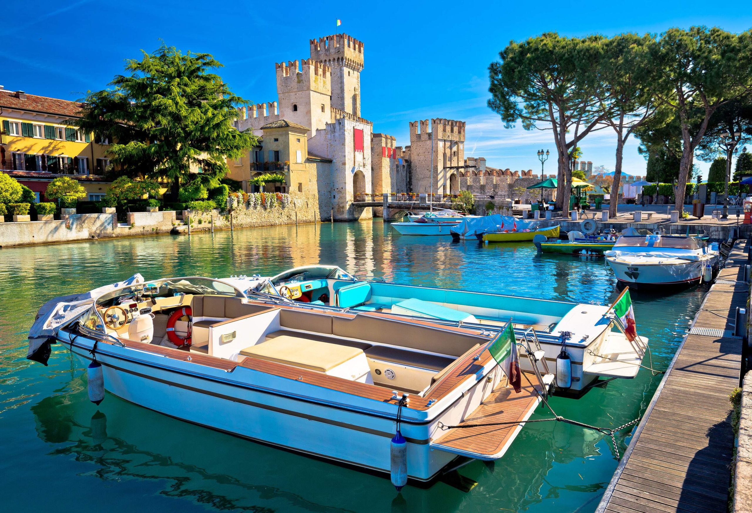 Yachts anchored along the pier in a lake, with a Roman castle in the background.