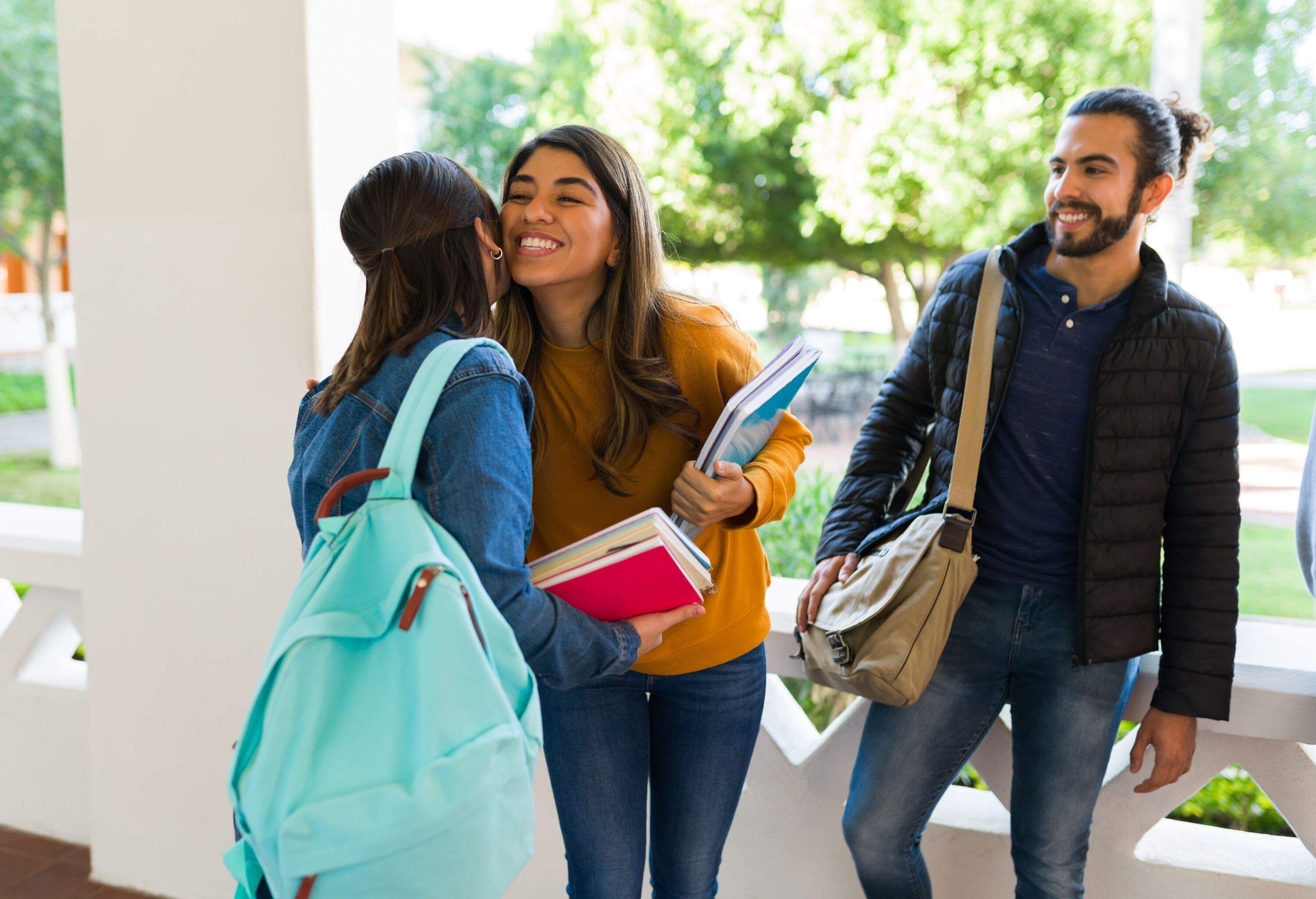 Happy young woman giving a kiss on the cheek to her best friend at the university while carrying books to class