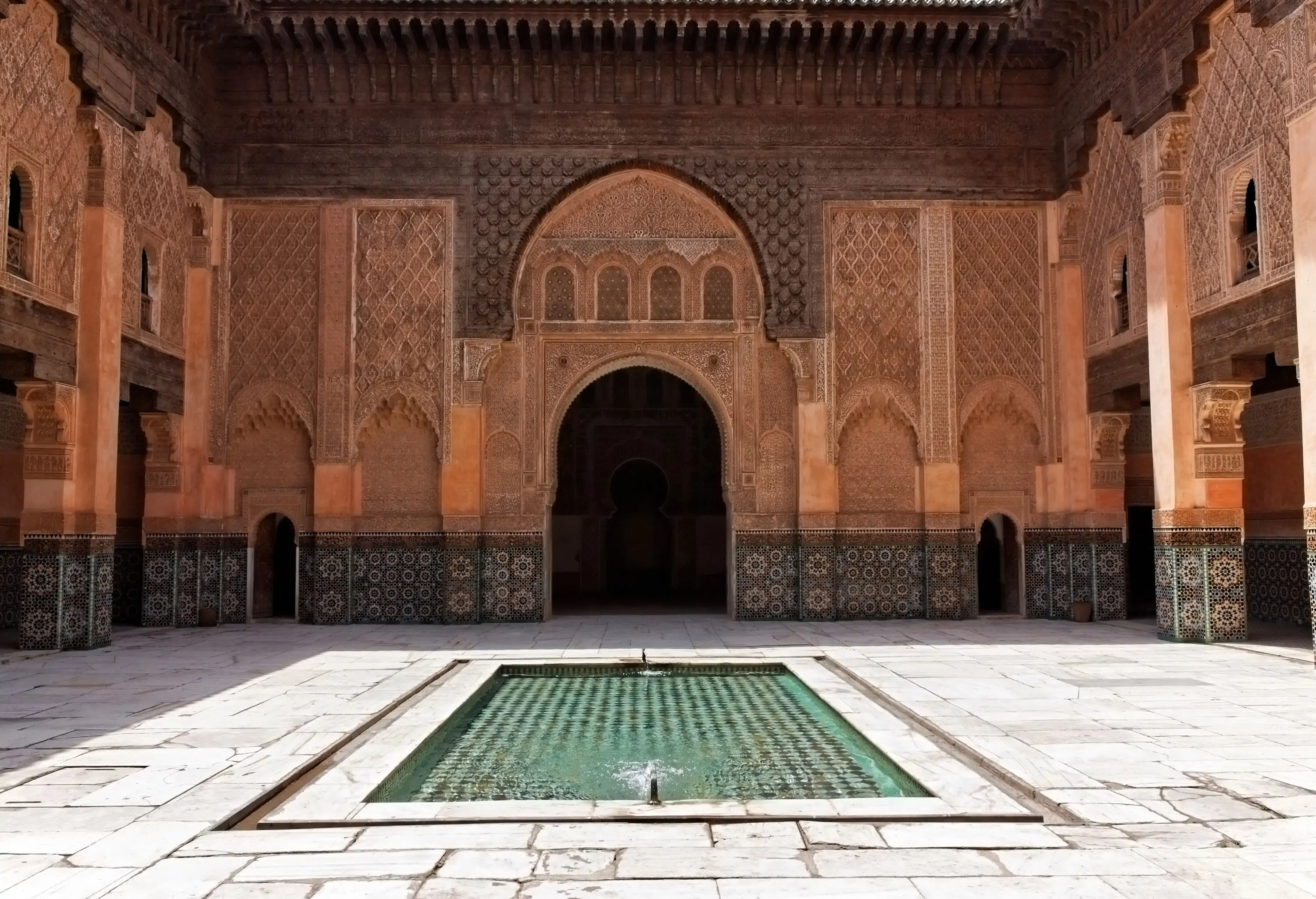 A reflective pool in the middle of a courtyard with carved walls and arched doorways.